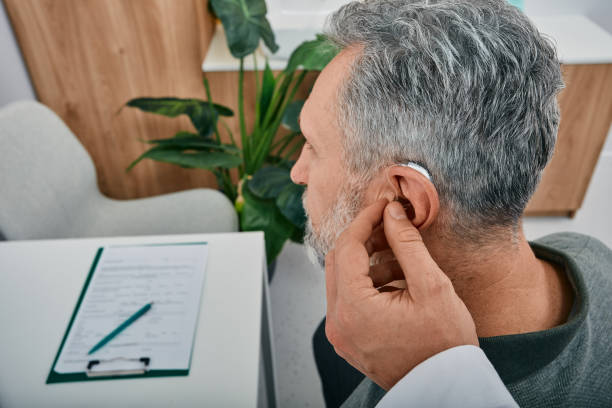 Audiologist fits a hearing aid on deafness mature man ear while visit a hearing clinic. Hearing solutions for older stock photo