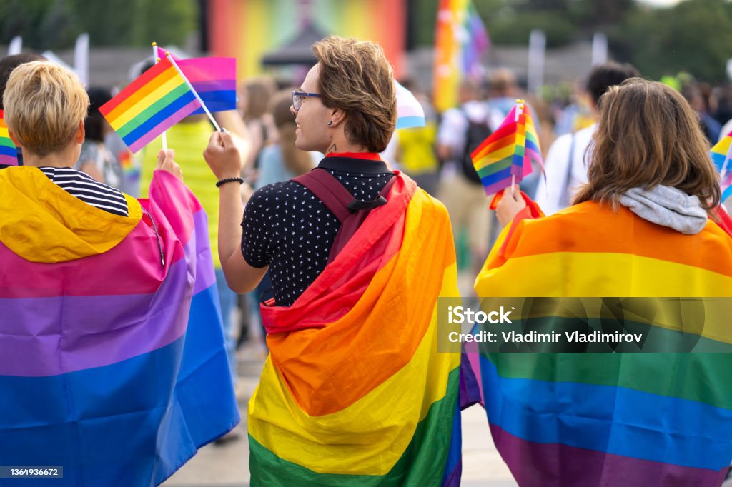 Group of people celebrating the pride month on a pride event Wrapped in bisexual flag and pride flags this trio are waving small pride flags and watching a gay pride event LGBTQIA Pride Event Stock Photo