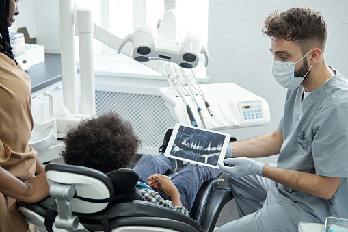 Dentist in protective workwear showing jaw x-ray on tablet screen to little patient sitting in armchair in dentistry office