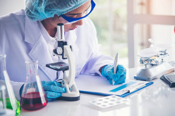 istock Woman scientist hand write note information in medical lab look at science microscope medical test, research biology chemistry. Asian Females technician laboratory analyzing blood test pharmaceutical 1364945965