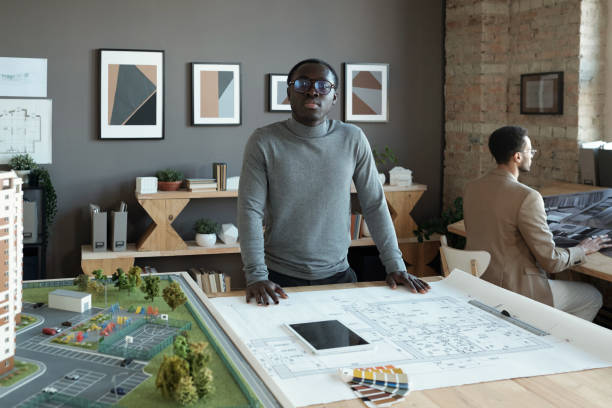 istock Young African engineer standing by table with sketch and house layout 1364947214