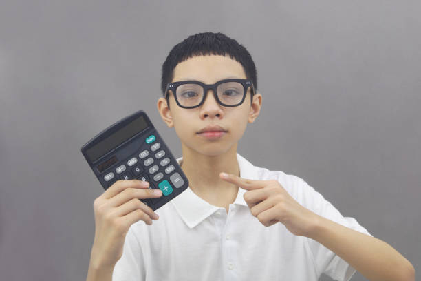 Friendly Asian boy in glasses holding calculator on gray background.