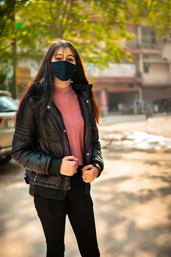 Outdoor portrait of beautiful, fashionable Indian young woman in black warm jacket and protective face mask in winter. She is standing on road and looking at the camera.