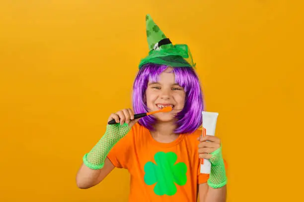 St.Patrick 's Day. Child girl a carnival costume leprechaun green wig, hat holds with toothpaste and an orange brush isolated on a yellow background. Medicine, dental hygiene, holidays concept.