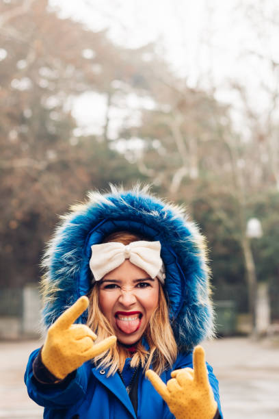 jeune femme moderne, vêtue d’un manteau bleu, de gants jaunes et de rouge à lèvres, en hiver, faisant symboliser les cornes de métal lourd et tirant la langue devant la caméra. - blue hair winter women cold photos et images de collection