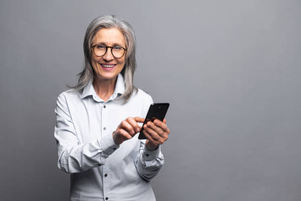 glückliche geschäftsfrau im formellen hemd, die den smartphone-bildschirm berührt, während sie sich mit den neuen telefonfunktionen großartig fühlt. indoor-studio isoliert auf grau aufgenommen - message holder stock-fotos und bilder