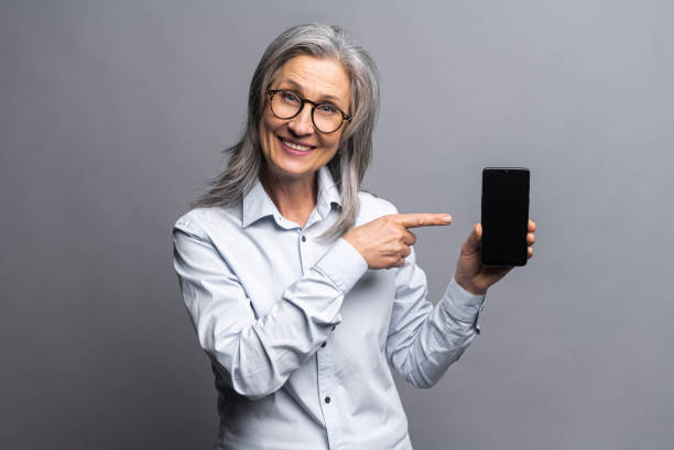 reife grauhaarige geschäftsfrau in formeller kleidung, die smartphone zeigt und mit einem finger auf den bildschirm zeigt, mit leerem platz für werbung, isoliert stehend - message holder stock-fotos und bilder