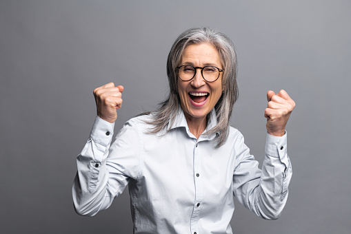 Portrait of excited overjoyed businesswoman standing with raised fists and shouting yeah, I'm winner, rejoicing something, success. Studio shot isolated on grey background