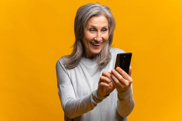 istock Shocked senior woman standing, holding and pointing at mobile display, screen with surprised face. Indoor studio shot, isolated on yellow 1364982064