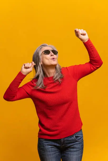 Mature lady in sunglasses dancing active isolated on yellow. Joyful modern senior woman with gray-hair enjoying celebrating party