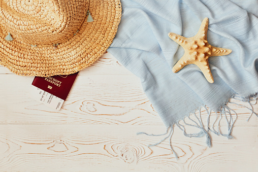 Women's straw hat, red passport and plane ticket, blue tippet and starfish on a white wooden background - summer vacation concept. Flat lay