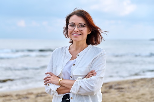 Outdoor portrait of a successful middle aged woman with folded hands. Confident mature female in glasses looking at the camera, gray cloudy sky sea background