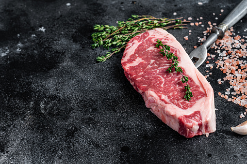 Raw striploin steak on a butcher table with salt and thyme. Black background. Top view. Copy space.