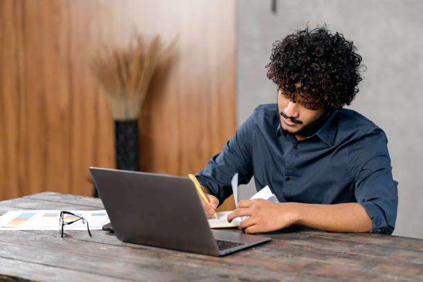 istock Online training. Young guy student with curly hair studying at home while sitting in the modern room and using laptop. He writing in a notebook 1364993068
