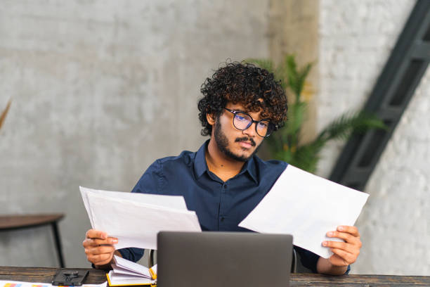 Executive mixed-race male office employee doing paperwork while sitting at the desk with laptop in office. Focused and serious Hindu man looking through sheets of documents