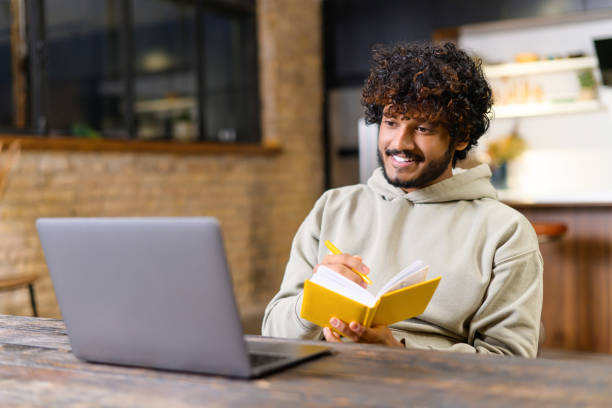 istock Handsome young man in smart casual wear is using laptop computer for watching online classes and webinars. He is taking notes at notebook, while sitting at home office 1364994259