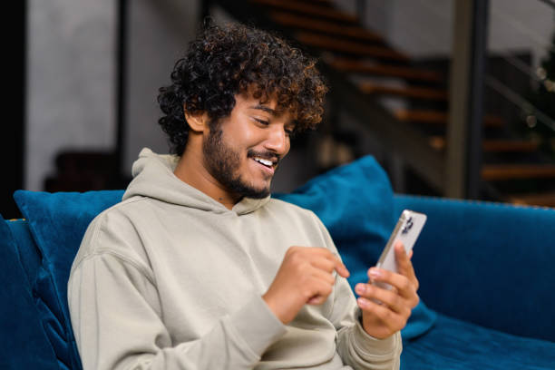 istock Focused bearded Indian man sitting on the sofa at home and holding smartphone. Hispanic guy reading news online, spend leisure time in social networks 1364994576