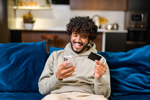 Smiling Indian man holding banking card and smartphone, making purchases or ordering online. Mixed-race guy using mobile app for booking or renting, paying online with credit card sitting on the coach