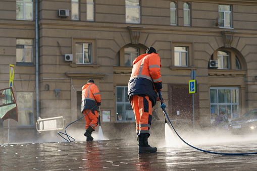Moscow, Russia - March 25, 2020:; Using water. Worker watering from a hose outdoor. Jet spray of water in sunlight, Concept of freshness, street cleaning in Coronavirus Pandemic reality.