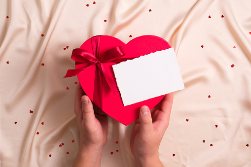 Man's hands hold a heart-shaped box with candy and a blank card over a silk background. Happy Valentine's Day concept