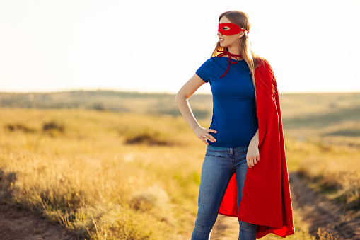 Joyful beautiful woman in a superhero costume posing against the backdrop of a sunset in nature. Superhero woman. Young and beautiful blonde in the image of a superheroine with a red cloak