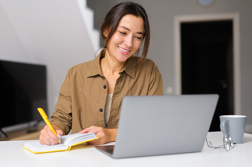 istock Smiling kind young woman works from home. She sitting at the kitchen table In front of the laptop and notebook 1365001960