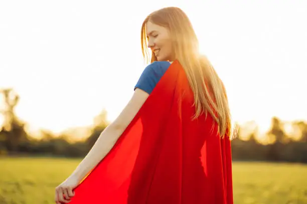Photo from the back Woman superhero. blonde in the image of a superheroine in a red cloak is growing up. Joyful beautiful woman in superhero costume posing against sunset background outdoors