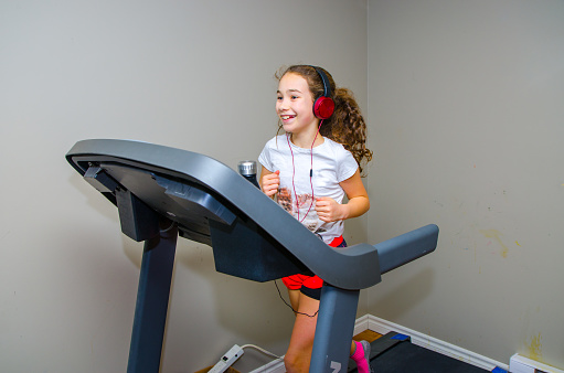 Girl jogging on home exercise treadmill in the basement without running shoes, wearing socks, shorts, t-shirt and pony tail. She's listening music with headphones. She's smiling.