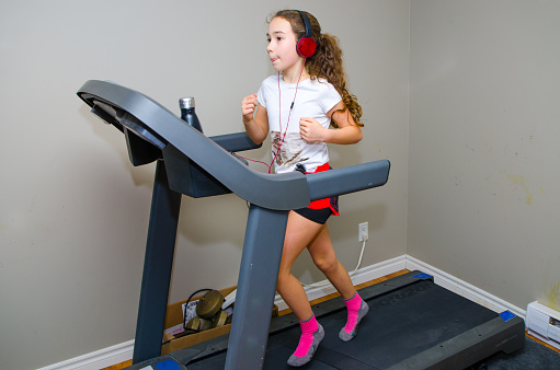 Girl jogging on home exercise treadmill in the basement without running shoes, wearing socks, shorts, t-shirt and pony tail. She's listening music with headphones. She has her tongue out.