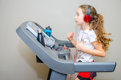 Girl jogging on home exercise treadmill in the basement without running shoes, wearing socks, shorts, t-shirt and pony tail. She's listening music with headphones.