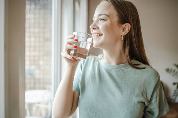 istock Young woman drinks water from a glass by the window 1365015579
