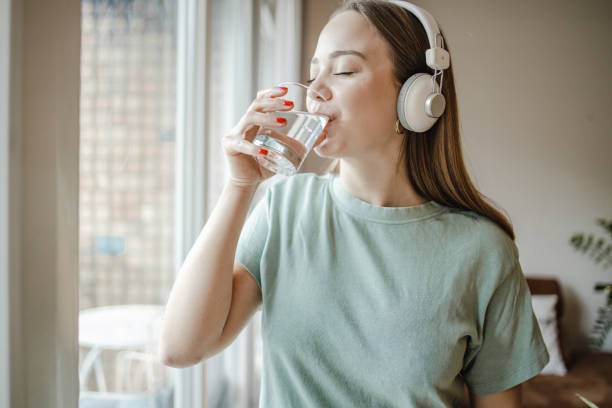istock Young woman drinks water from a glass by the window 1365015663