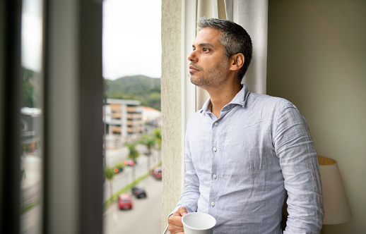 Man drinking a cup of coffee and looking lost in thought while looking out at the city through a window at home