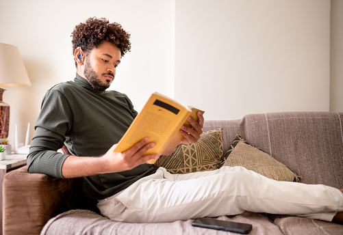 Young man with a hearing aid reading a book while relaxing on his living room sofa at home