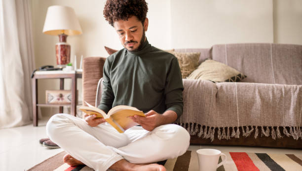 istock Young man reading a book on his living room floor 1365018176