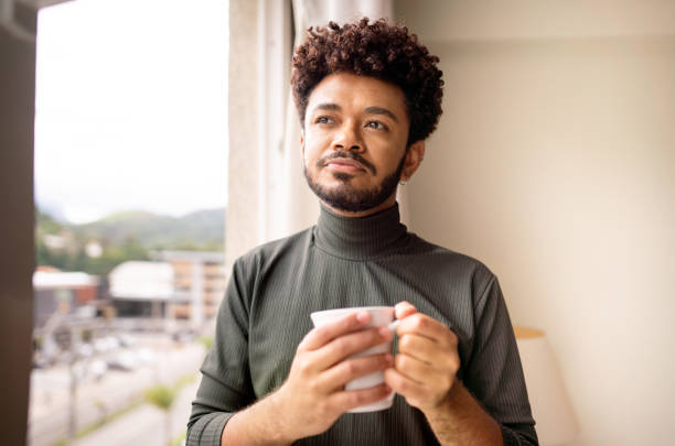 istock Young man looking out of a window at home while drinking coffee 1365018243