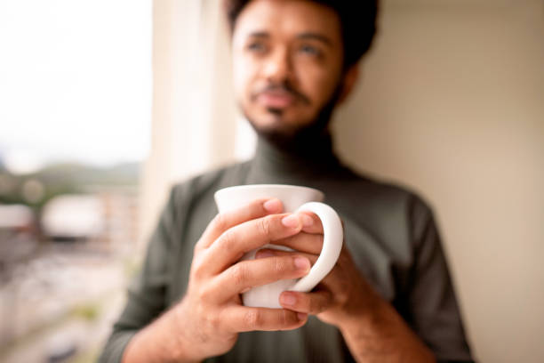 jeune homme buvant du café près d’une fenêtre à la maison - homme mug regarder dehors photos et images de collection
