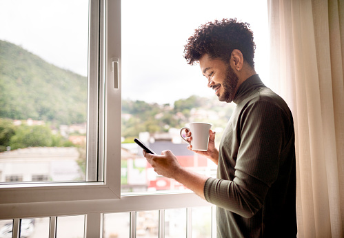 Smiling young man with adult braces standing by a window at home and checking his phone over a cup of coffee
