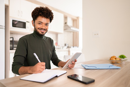 Portrait of a smiling young man with a hearing aid writing notes while working online at a table at home using a digital tablet