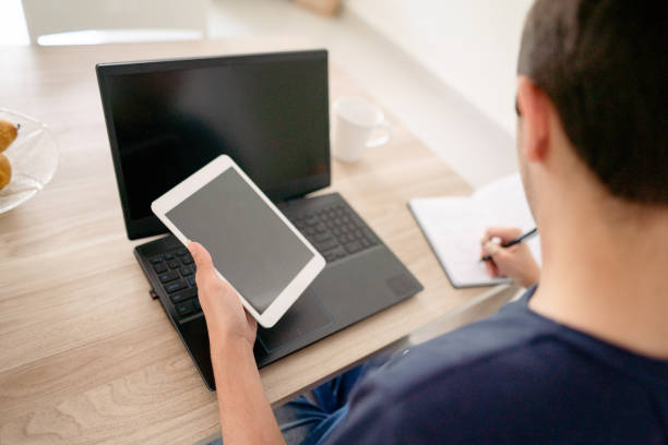 Teenage boy writing notes while doing homework with a digital tablet and a laptop at a table in his kitchen at home