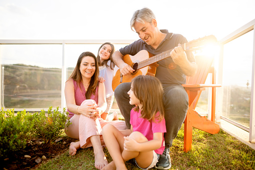 Dad playing guitar while sitting with his wife and two cute little daughters outside in their back yard on sunny afternoon