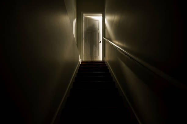 階段の上にわずかに開いたドアで照らされた暗い階段。 長い露出で撮影し、階段の上部にある姿のような幽霊のシルフエットの効果を生み出します。 - spooky ストックフォトと画像