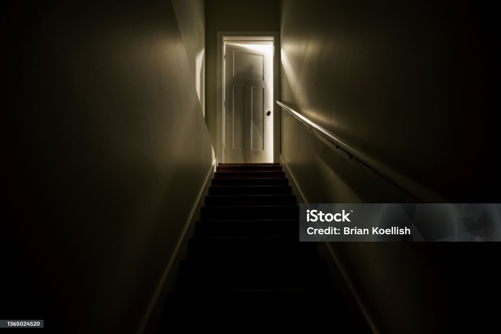 A dark stairwell illuminated by a slightly opened door at the top of the stairs.  Shot with a long exposure to create the effect of a sillhouette of a ghost like figure at the top of the stairwell. Spooky Stock Photo