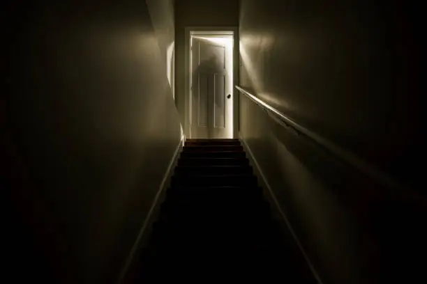 Photo of A dark stairwell illuminated by a slightly opened door at the top of the stairs.  Shot with a long exposure to create the effect of a sillhouette of a ghost like figure at the top of the stairwell.