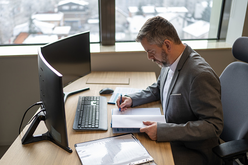 Side view of grey haired businessman in his office writing notes in a notepad at a desk.