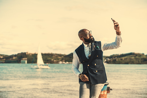 A dapper African-American senior in a fashionable outfit is sitting on a bollard next to the river and having video call on a dazzling day; an elegant black guy in a costume is taking a selfie on pier