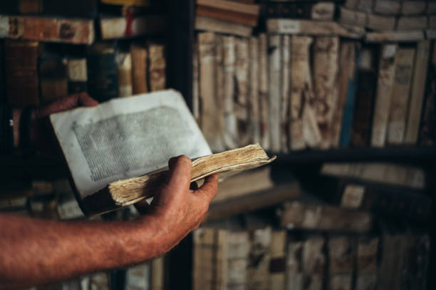 Color image of some old books piled on a wood desk, with one open in foreground.