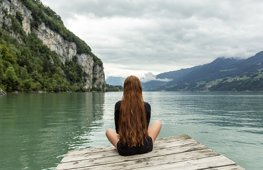 Calm girl sitting alone meditating on a wooden dock facing the beautiful lake and mountain view. Back view.