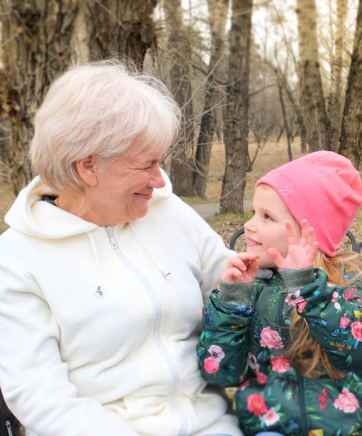 бабушка с внучкой обнимается и разговаривает, сидя на скамейке в парке - senior adult winter senior women daughter стоковые фото и изображения