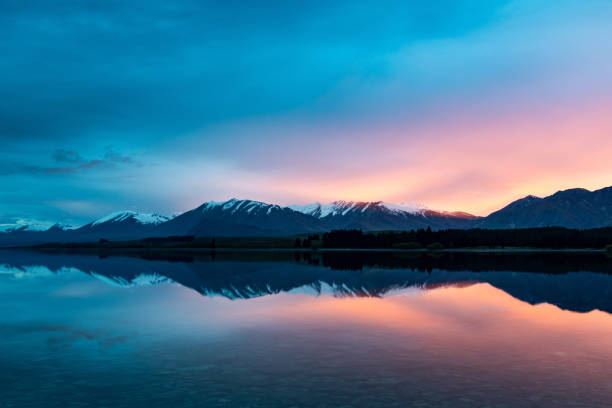 This October 2021 dawn image shows the glow from the sunrise through clouds at Lake Tekapo, Aotearoa New Zealand. The surface of the lake is mostly calm and provides a strong reflection. The snow-capped mountains east of the lake are visible, including from left: Round Hill, Mount Ardmore, and Mount Hay. The oranges and yellows of this image were only visible for a few minutes because of the shifting clouds.